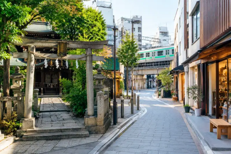 Tokyo’da geleneksel bir tapınak girişi, modern dükkânlar ve tren hattının bir arada görüldüğü semt sokağı.